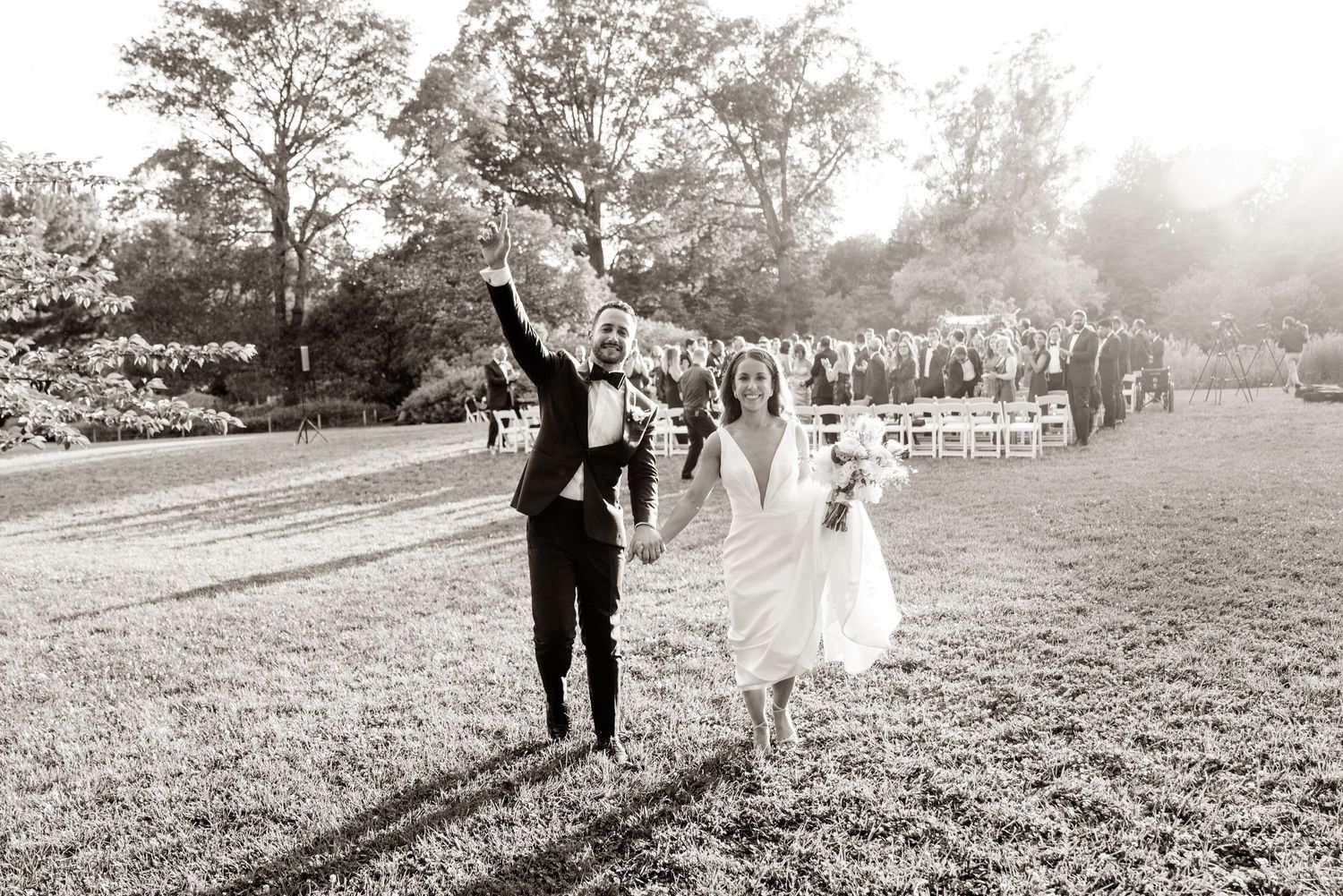 Black and white photo of joyful newlyweds running through field with wedding guests following behind.