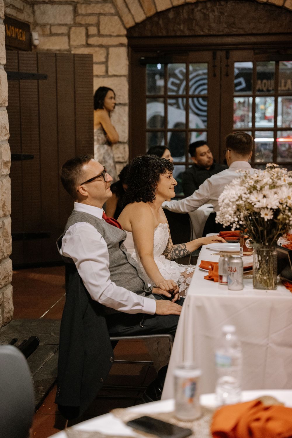 Wedding guests sit at round tables decorated with floral centerpieces during an indoor reception celebration.