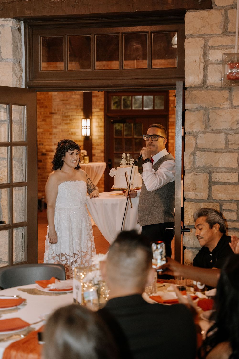 Wedding guests enjoy dinner and conversation at tables in a stone-walled reception venue.