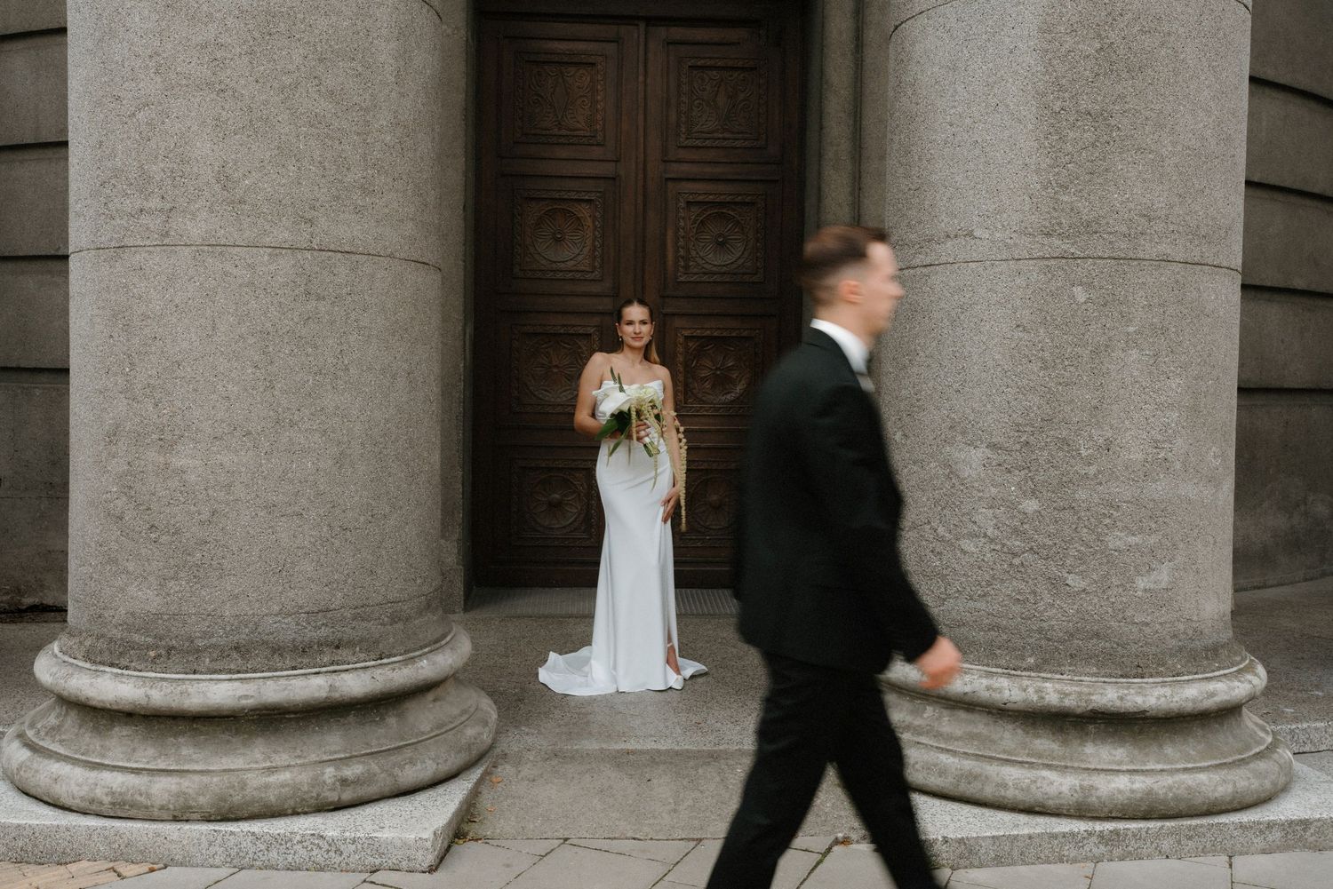 Elegant wedding scene with bride between classic stone columns as groom walks past.