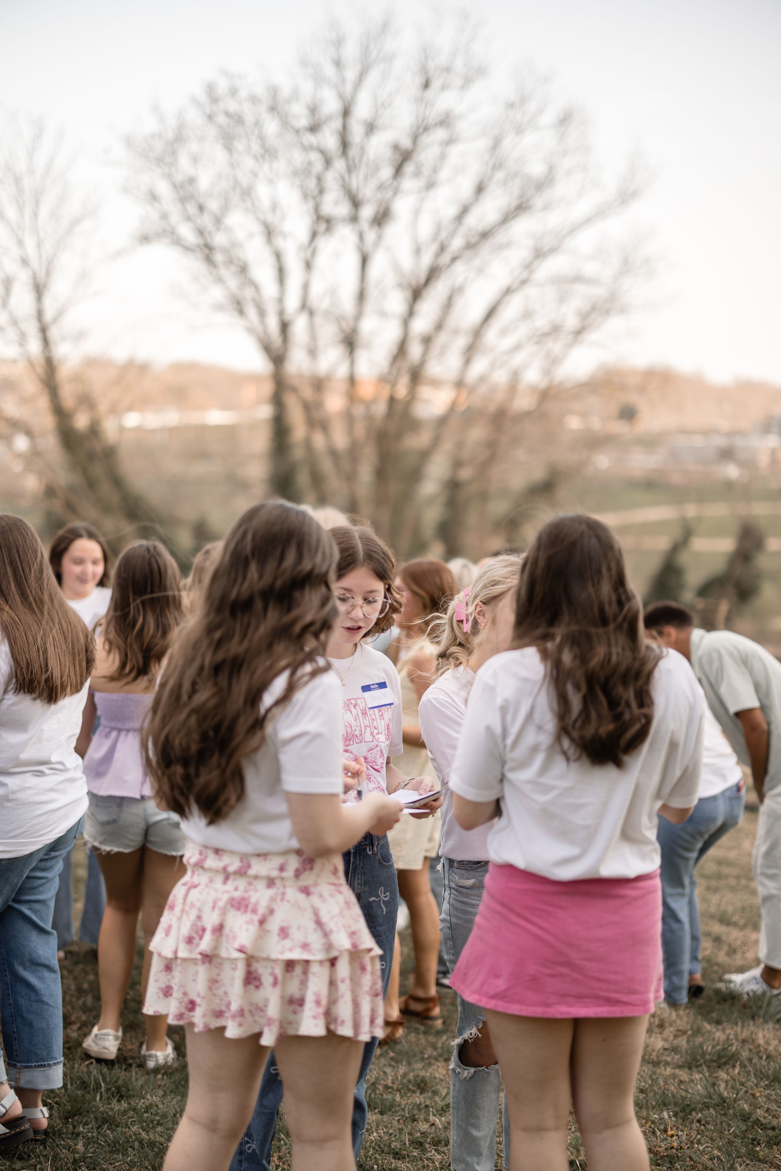 A group of students in white tops and casual attire gather outdoors on grass during sunset.