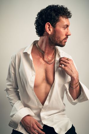 Bearded man with curly hair in an open white shirt, wearing layered gold chains, posing against a neutral background.