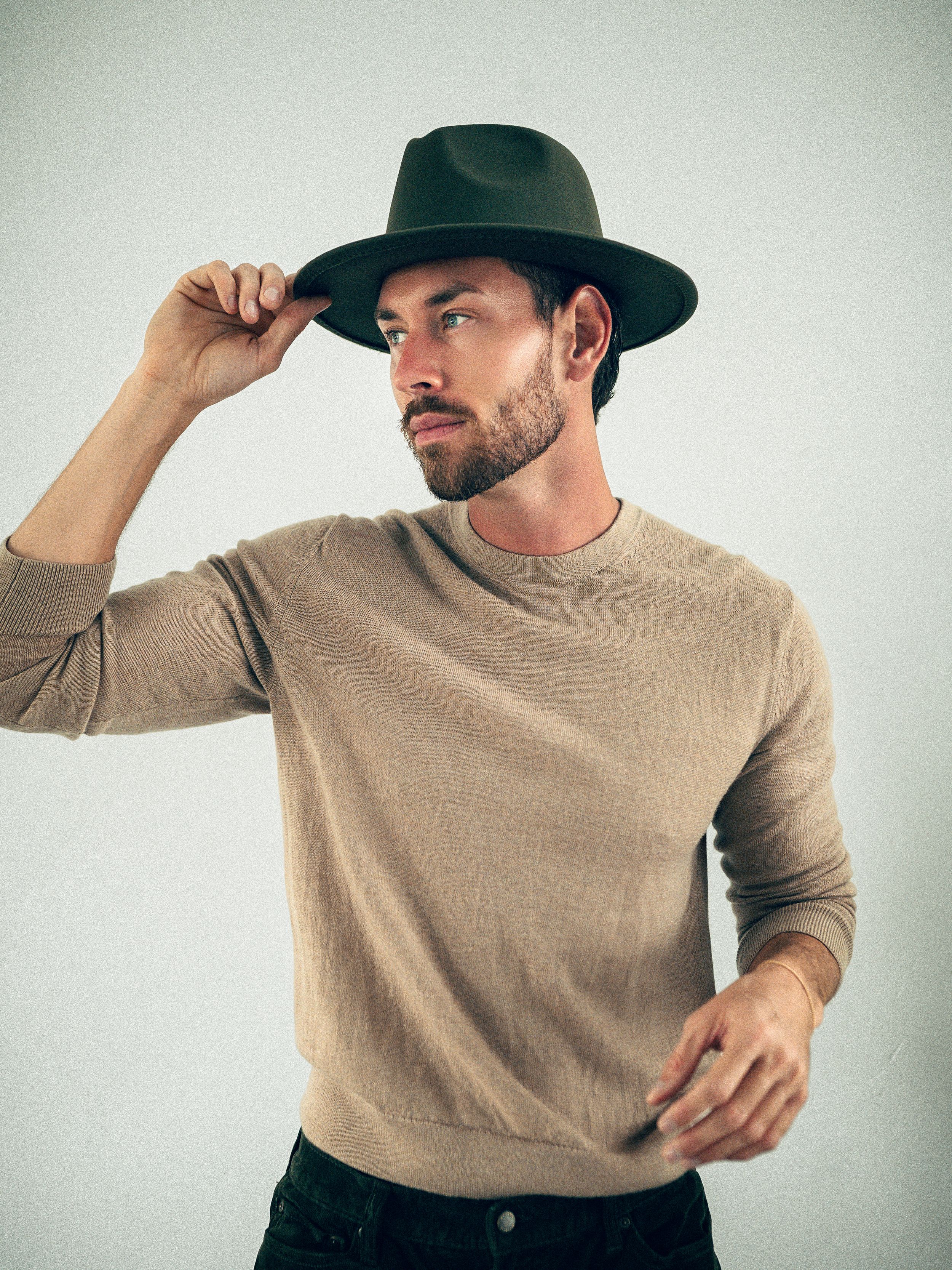 Stylish bearded man in a beige sweater adjusting a dark wide-brim felt hat against a light gray background.