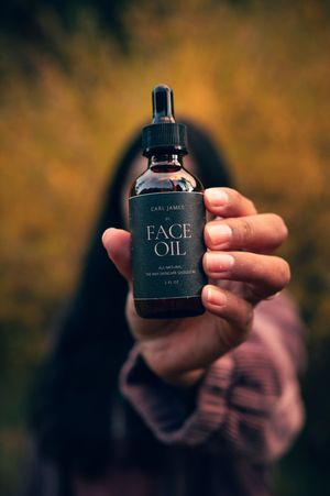 Woman with blurred face holding Carl James Face Oil bottle toward camera against golden autumn bokeh.