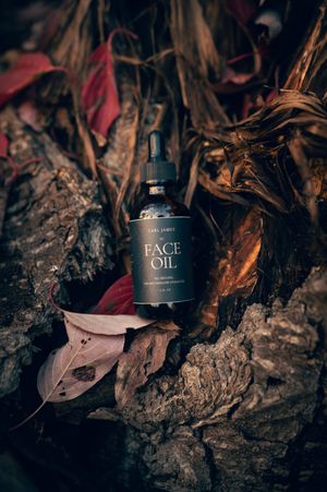 Carl James Face Oil dark glass bottle resting on rustic bark surrounded by dried autumn leaves.