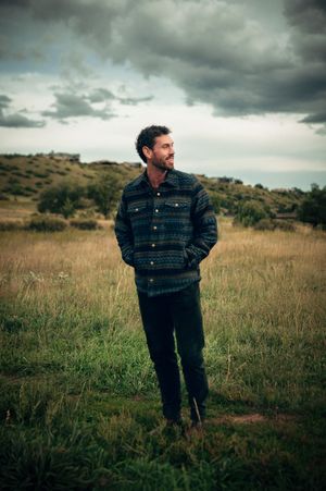Bearded man in patterned flannel shirt standing in grassy field under dramatic cloudy sky.