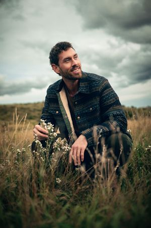 Smiling bearded man crouching in tall grass field holding wildflowers under overcast sky.