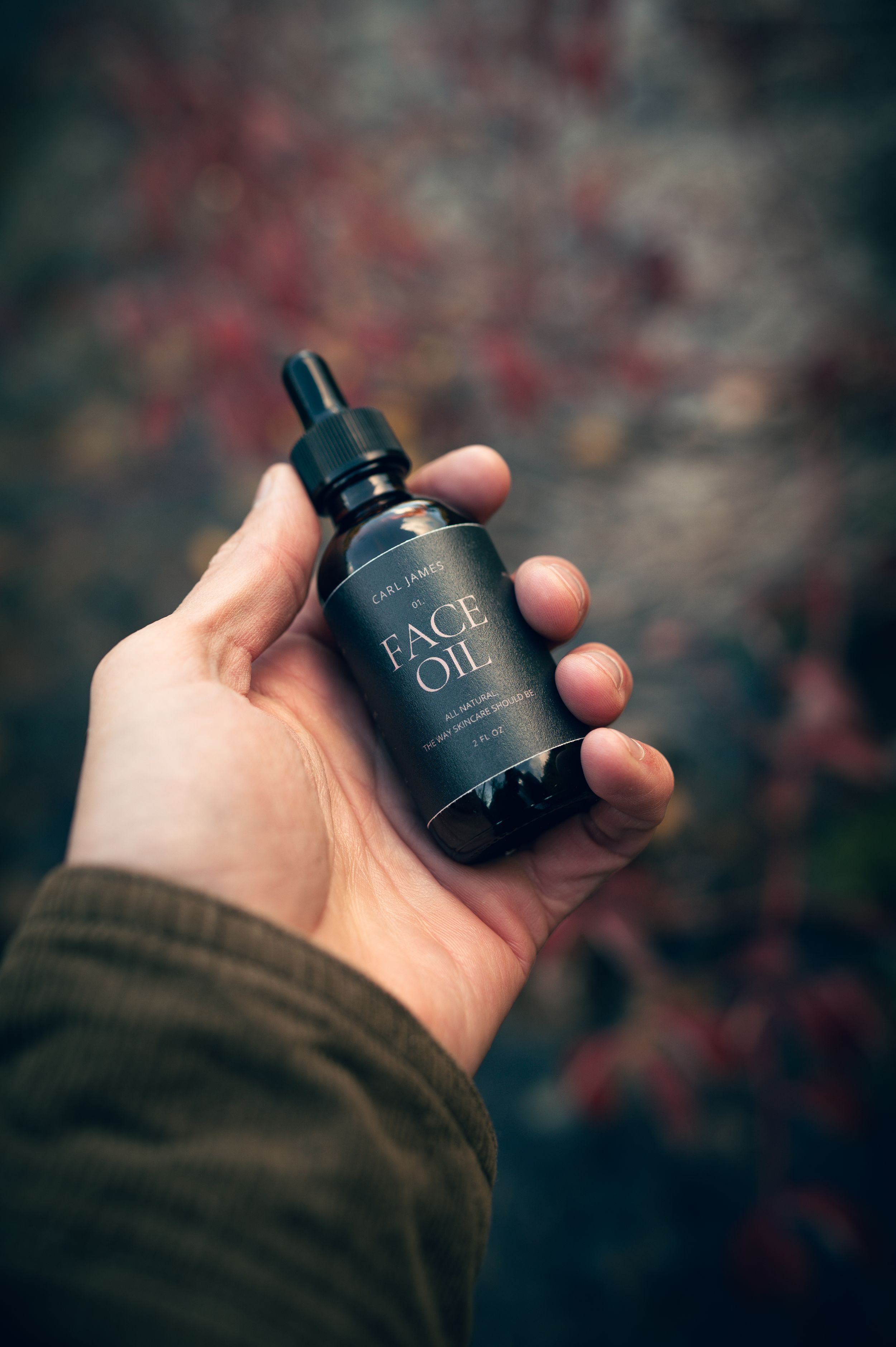 Hand holding dark amber dropper bottle of Carl James Face Oil with autumn foliage in background.