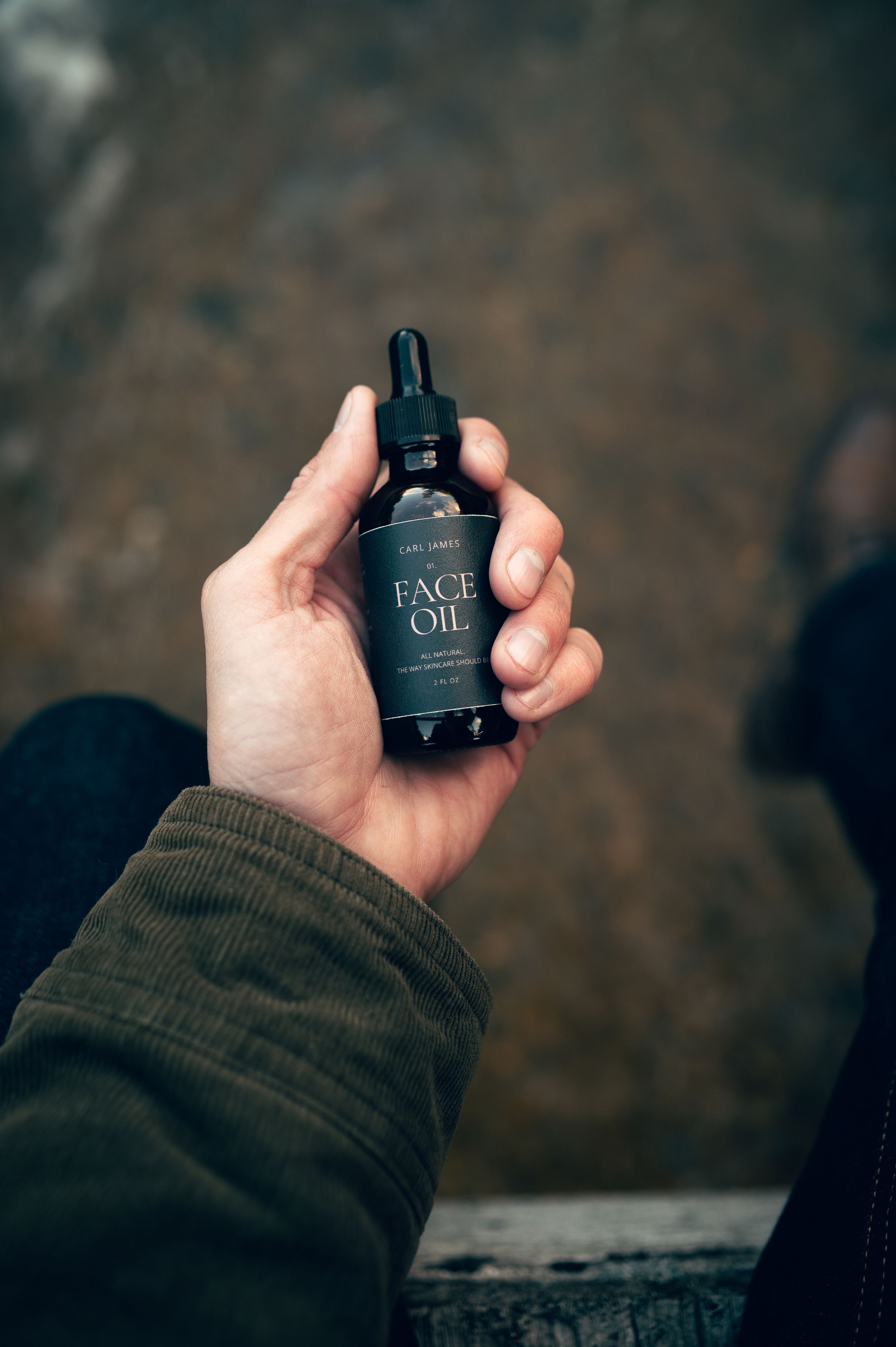 Close-up hand holding Carl James Face Oil dark glass dropper bottle outdoors on stone surface.