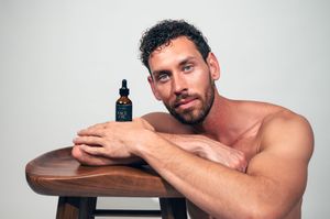 Shirtless bearded man resting chin on arms over wooden stool beside amber bottle of face oil.