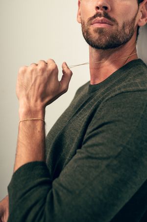 Close-up of bearded man in olive green sweater holding a thin gold chain necklace with gold bracelet.