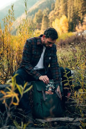 Bearded man in plaid shirt packing Lineage Classic meat sticks into green backpack in autumn wilderness.