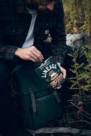 Man in flannel shirt opening Lineage Classic grass-fed beef meat sticks box beside green canvas backpack.