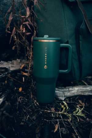 Forest green HydroJug tumbler with handle and gold accents resting on woodland ground near autumn foliage.