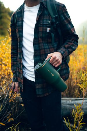 Man in plaid flannel shirt carrying a forest green HydroJug in a colorful autumn field landscape.