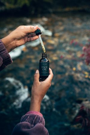 Hands holding Carl James Face Oil dropper bottle over a rocky stream with autumn leaves below.