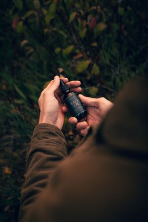 Person holding a dark glass bottle of Carl James Face Oil with dropper against green foliage background.