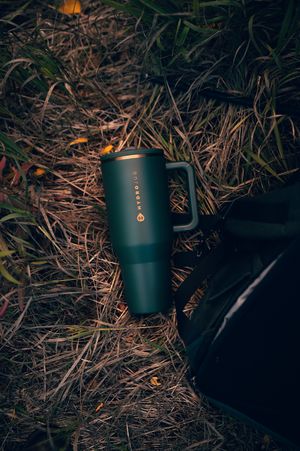 Dark green HydroJug insulated tumbler with handle resting on dry autumn grass next to black bag.