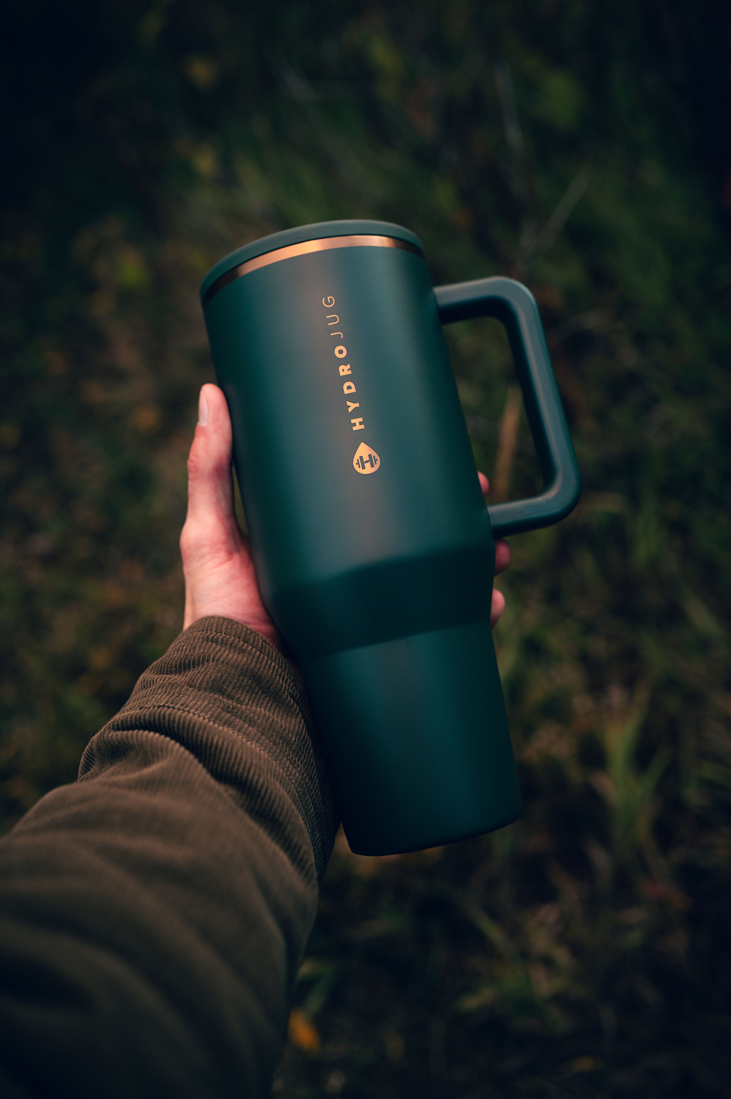 Hand holding dark green HydroJug tumbler with copper logo against blurred outdoor grass background.