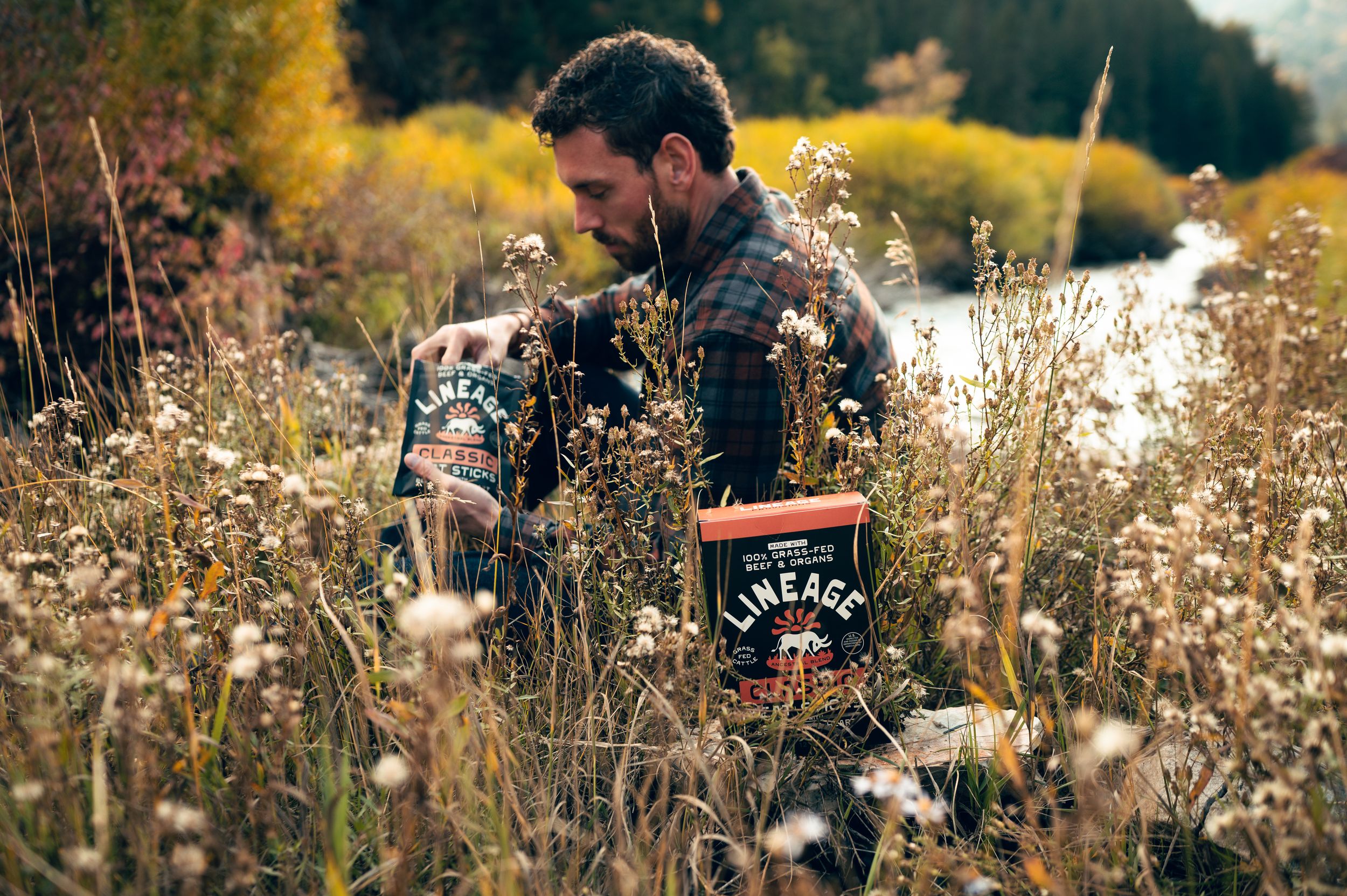 Man in flannel shirt holding Lineage Classic meat stick boxes while standing in golden autumn wildflower field.