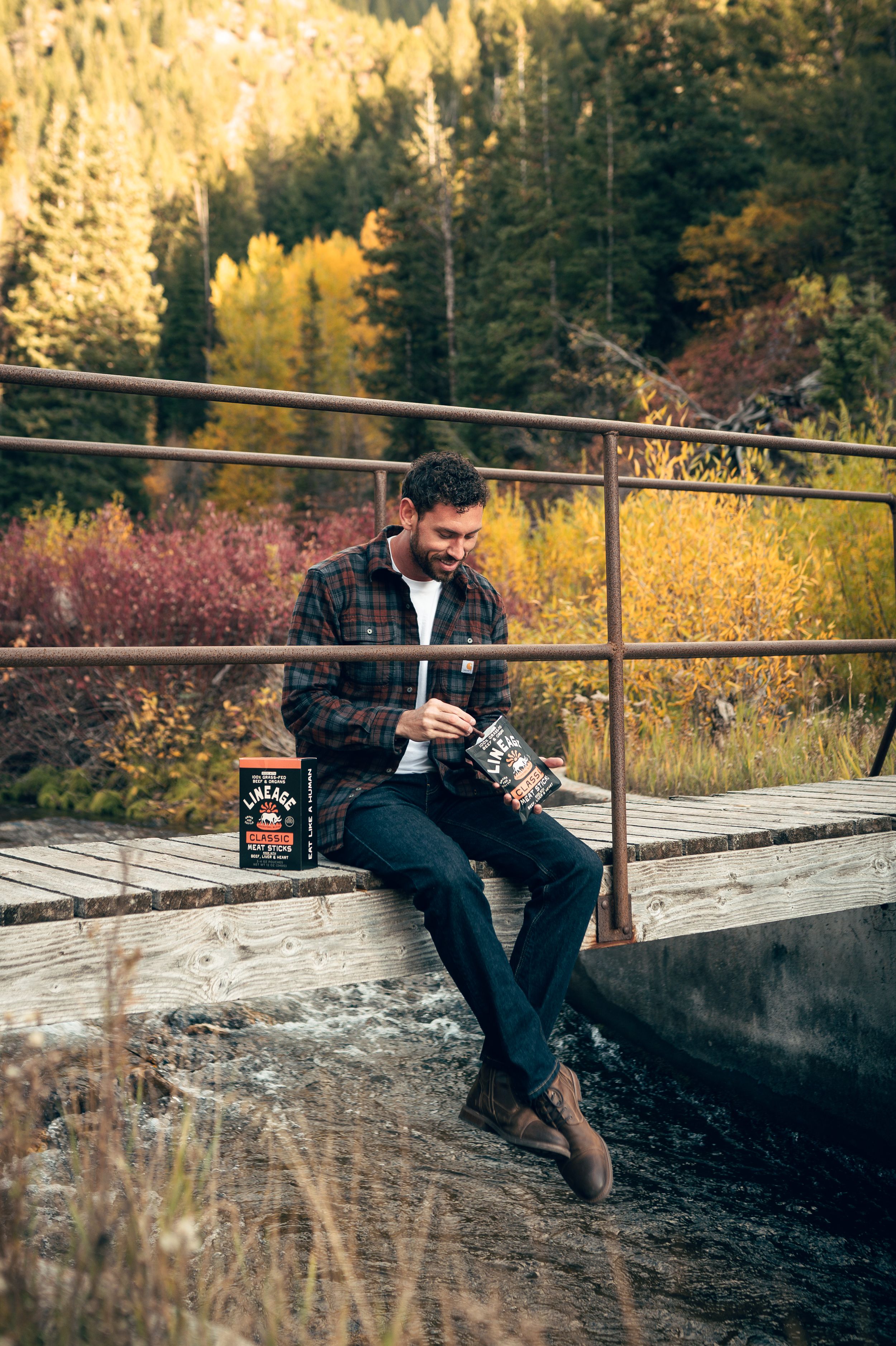 Bearded man in plaid shirt enjoying Lineage meat sticks on wooden bridge surrounded by colorful fall forest.