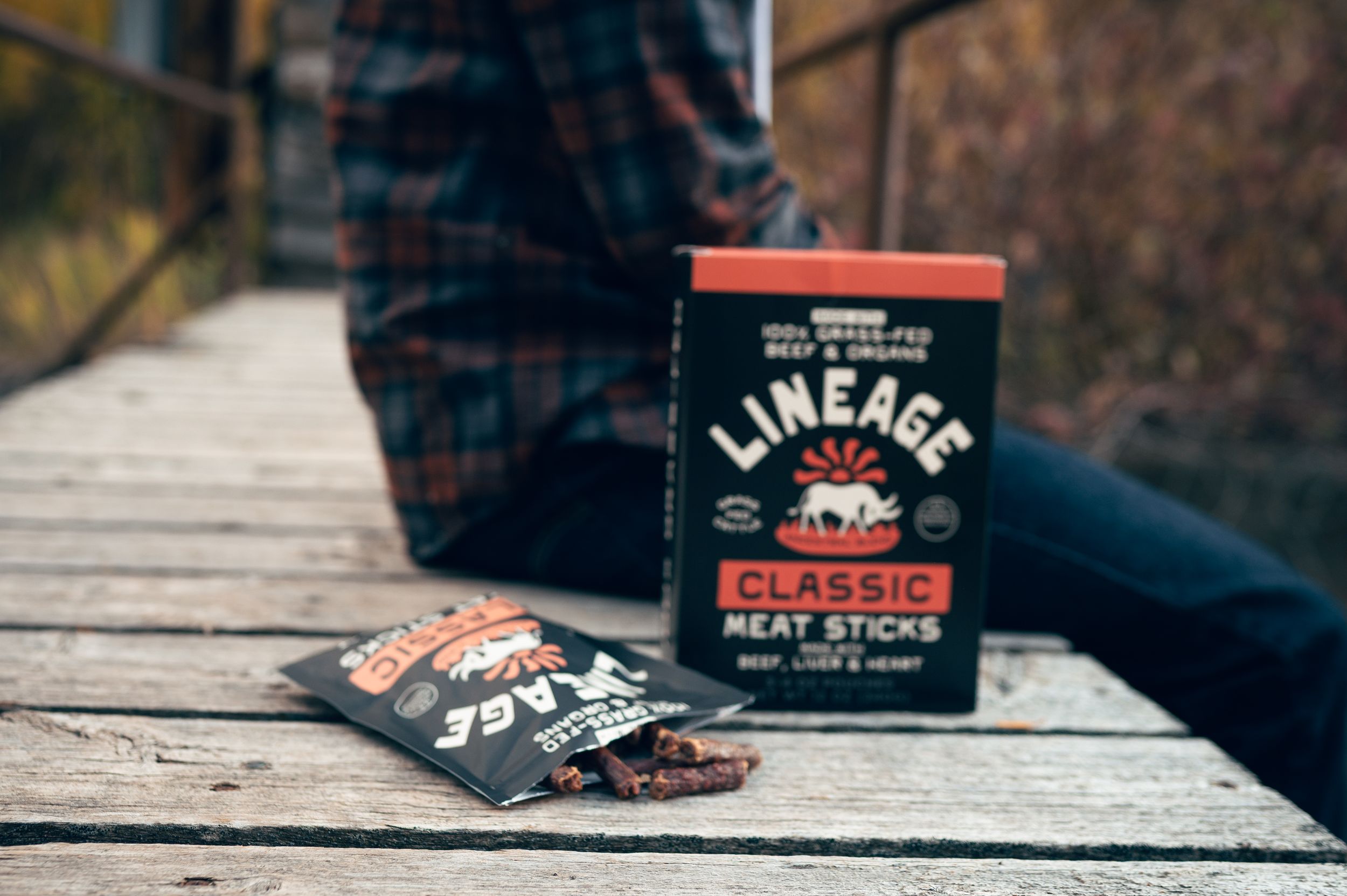 Lineage Classic grass-fed beef meat sticks box with open package on weathered wooden bridge planks.
