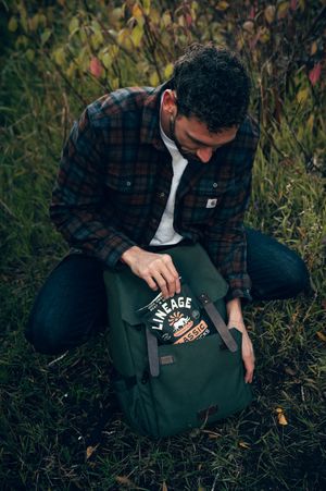 Curly-haired man in Carhartt flannel placing Lineage meat sticks into green canvas backpack outdoors.