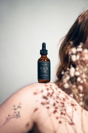 Amber dropper bottle of face oil balanced on bare shoulder with floral tattoo and dried flowers.