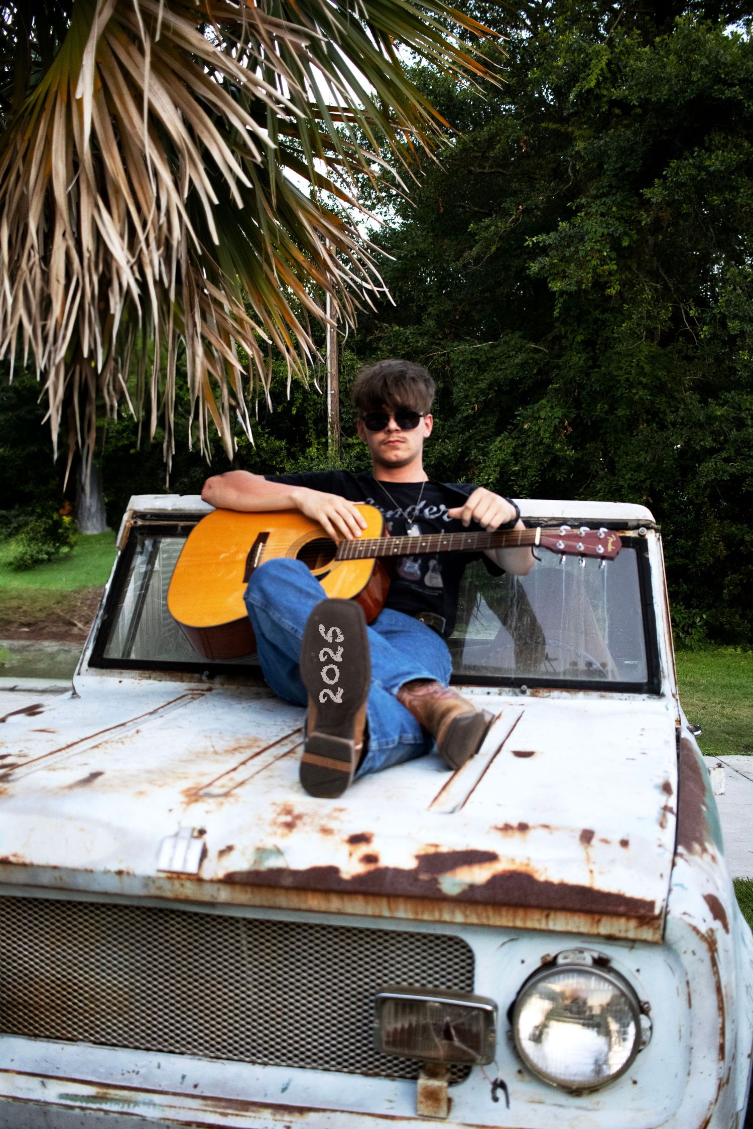Person playing guitar while sitting on rusty vintage truck hood under palm trees.