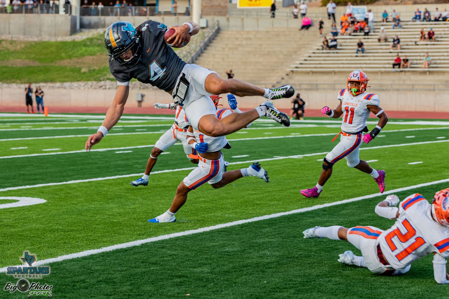 Football player leaps over defenders while carrying the ball during an intense game on artificial turf field.