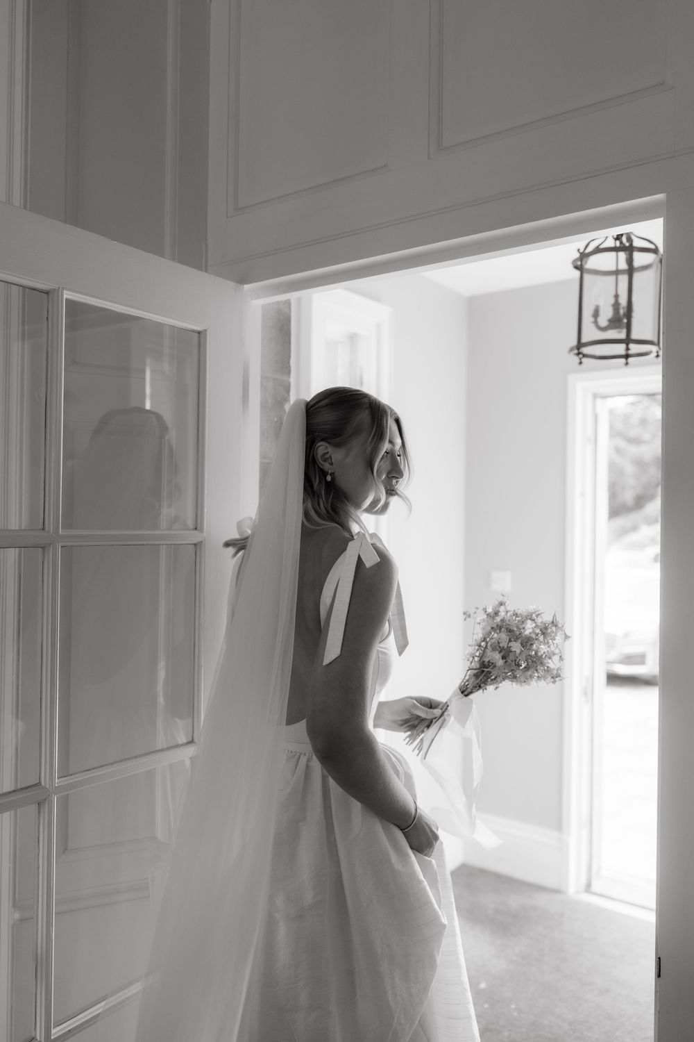 Black and white photo of a bride in a wedding dress holding a bouquet while standing in a doorway with natural light.