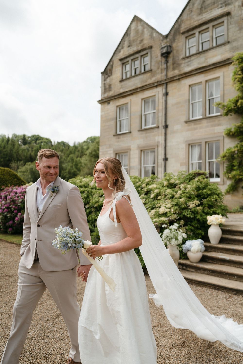 A wedding couple shares a joyful moment on the gravel path outside an elegant stone manor house.