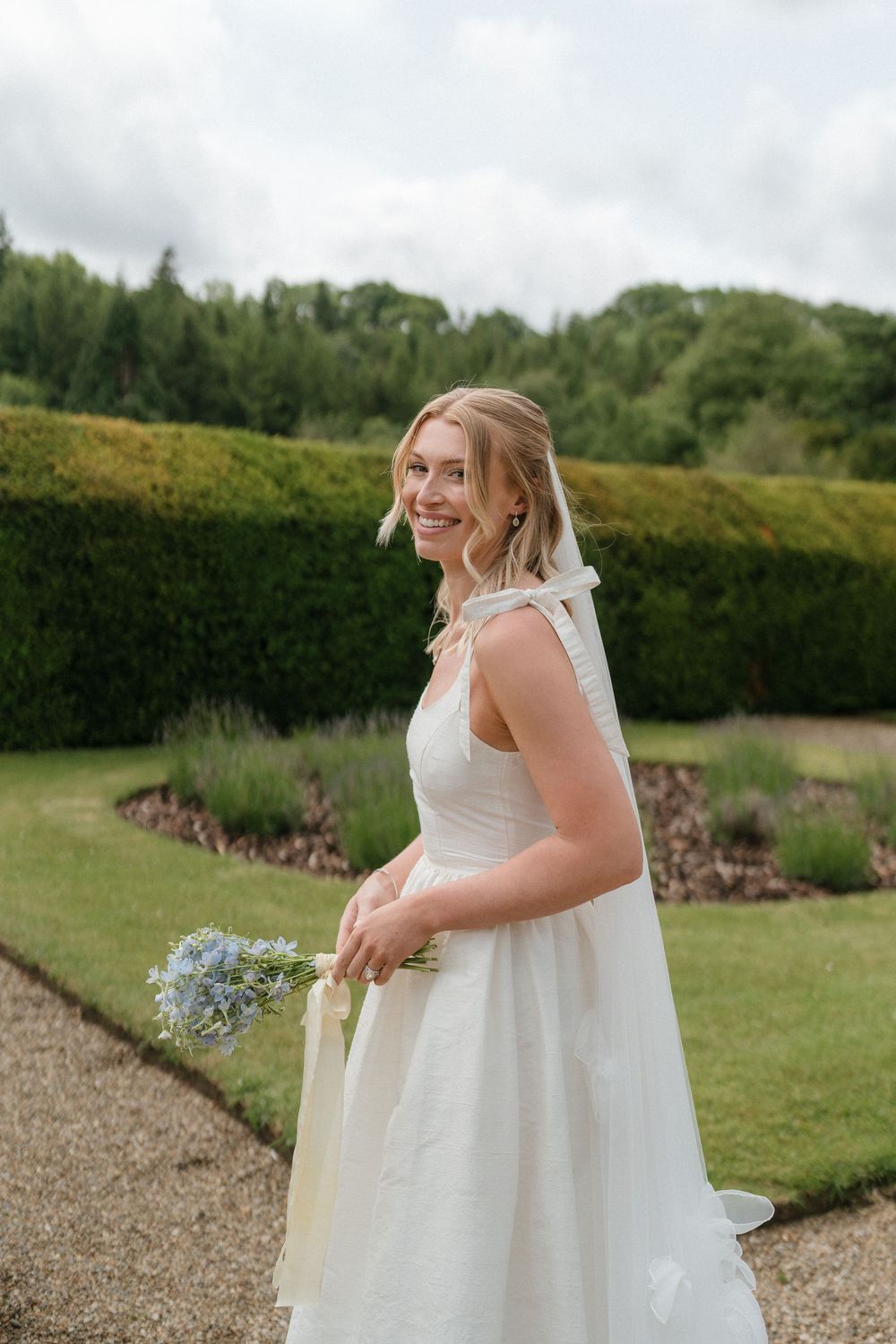 A person in a flowing white wedding dress stands in a garden setting with gravel paths and green hedges.