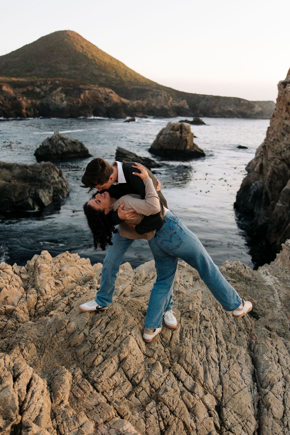 Adventurous couple posing on rocky coastal formations during sunset by the ocean.