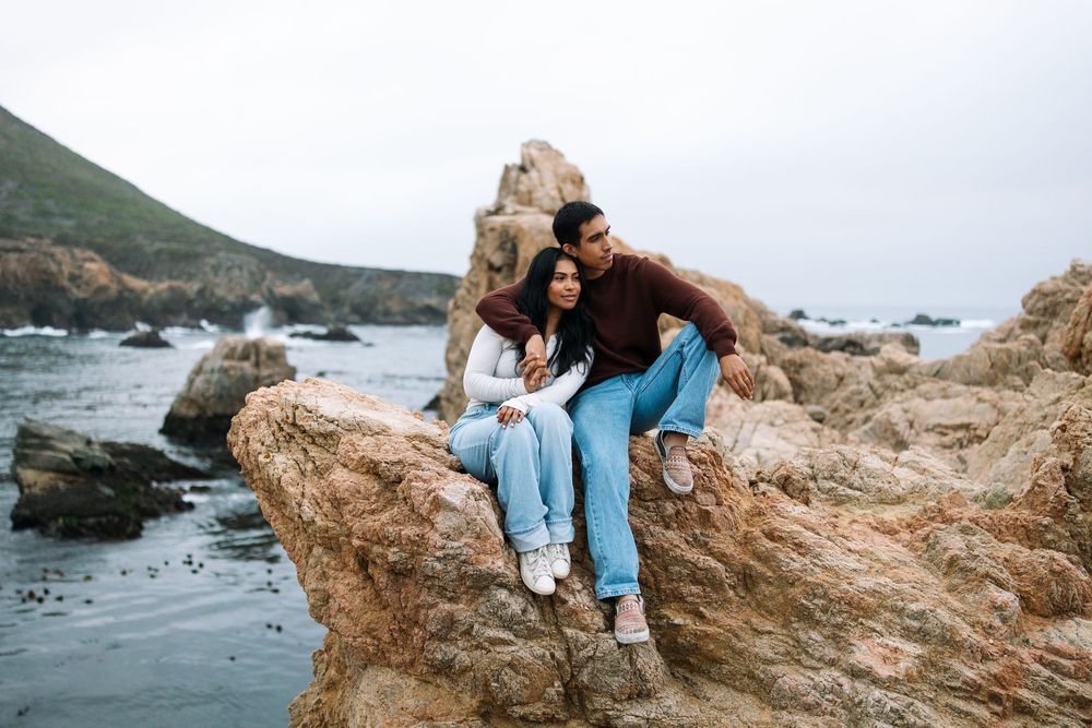 Couple sitting on coastal rocks overlooking the Pacific Ocean during a sunny afternoon.