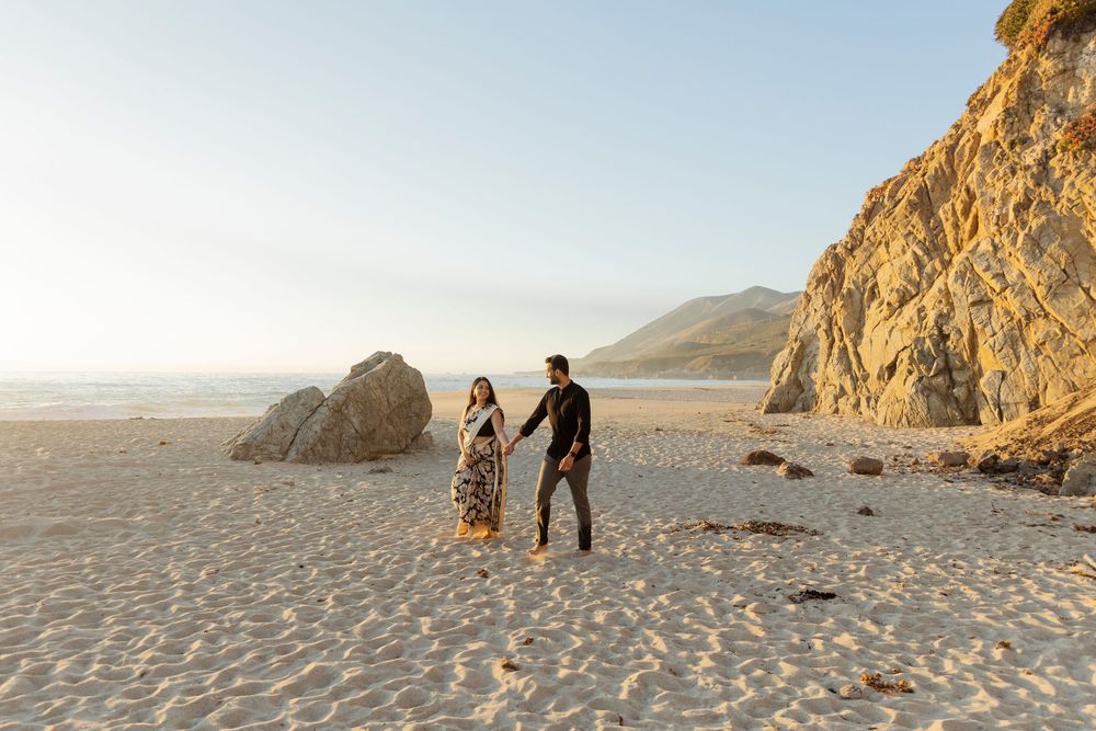 Couple walking along a sandy beach at sunset with rocky cliffs in the background.