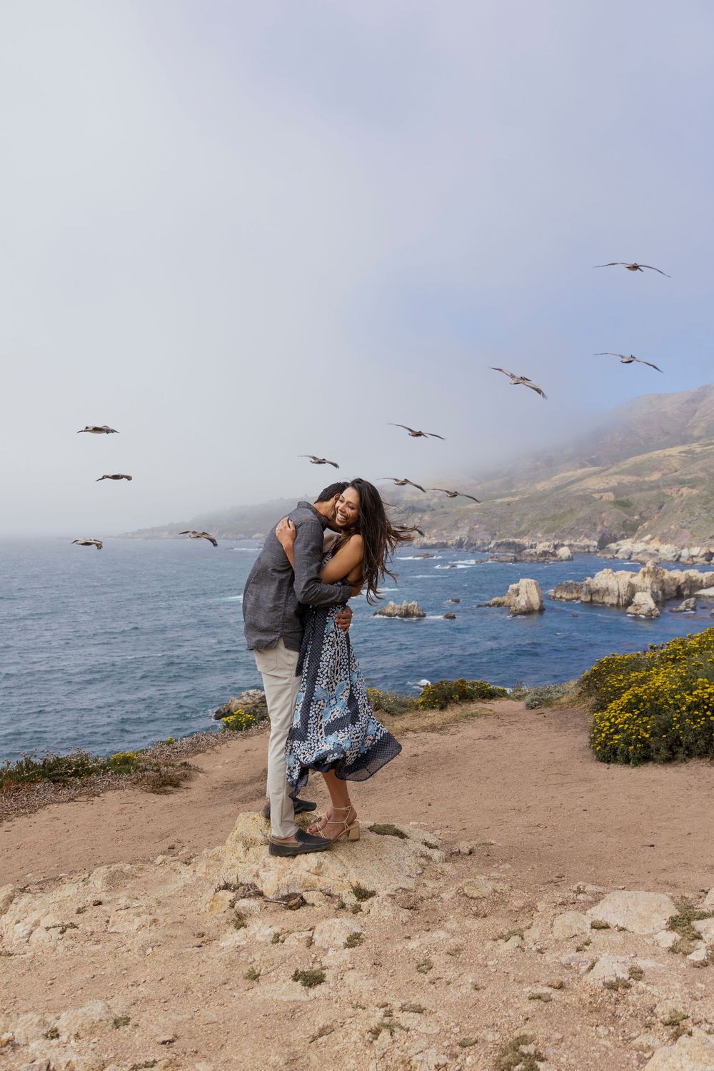 Playful coastal engagement photos with seagulls flying overhead at a scenic overlook.