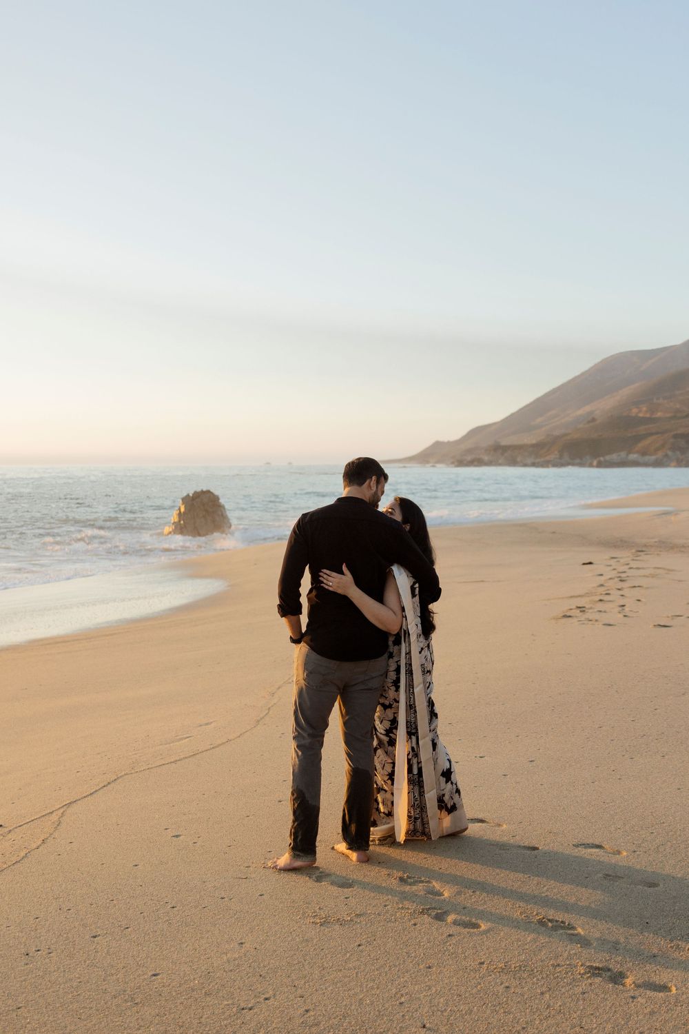 Romantic beach engagement session at sunset with couple silhouetted against sandy shoreline.