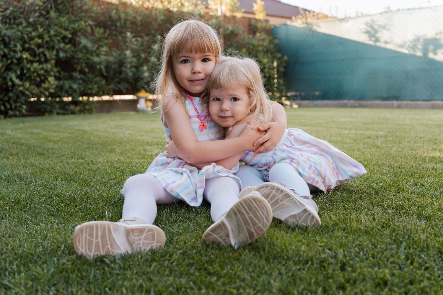 Two children in white-pink outfits sitti together on green grass during a sunny afternoon in Rouse Hill in garden during a family photoshoot