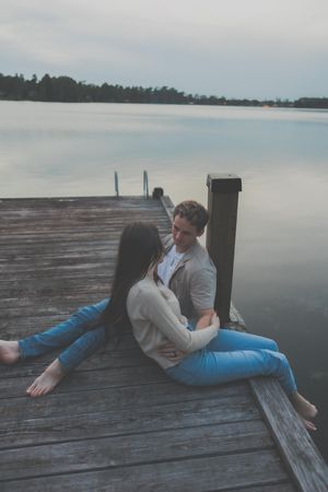 Two people lounging on a wooden dock overlooking a serene lake on an overcast day.