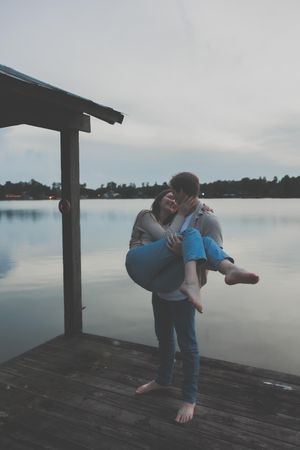 A romantic couple embraces on a wooden dock overlooking a misty lake during a moody, overcast day.