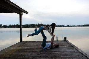 Two people perform an acrobatic yoga pose on a wooden dock overlooking a serene lake.