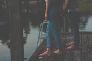 Couple's legs dangling over edge of wooden dock by calm water at sunset.