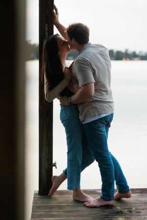 A romantic couple embraces while standing on a wooden dock by the water at sunset.