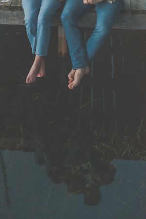 Bare feet dangle over the edge of a wooden dock above calm reflective water.