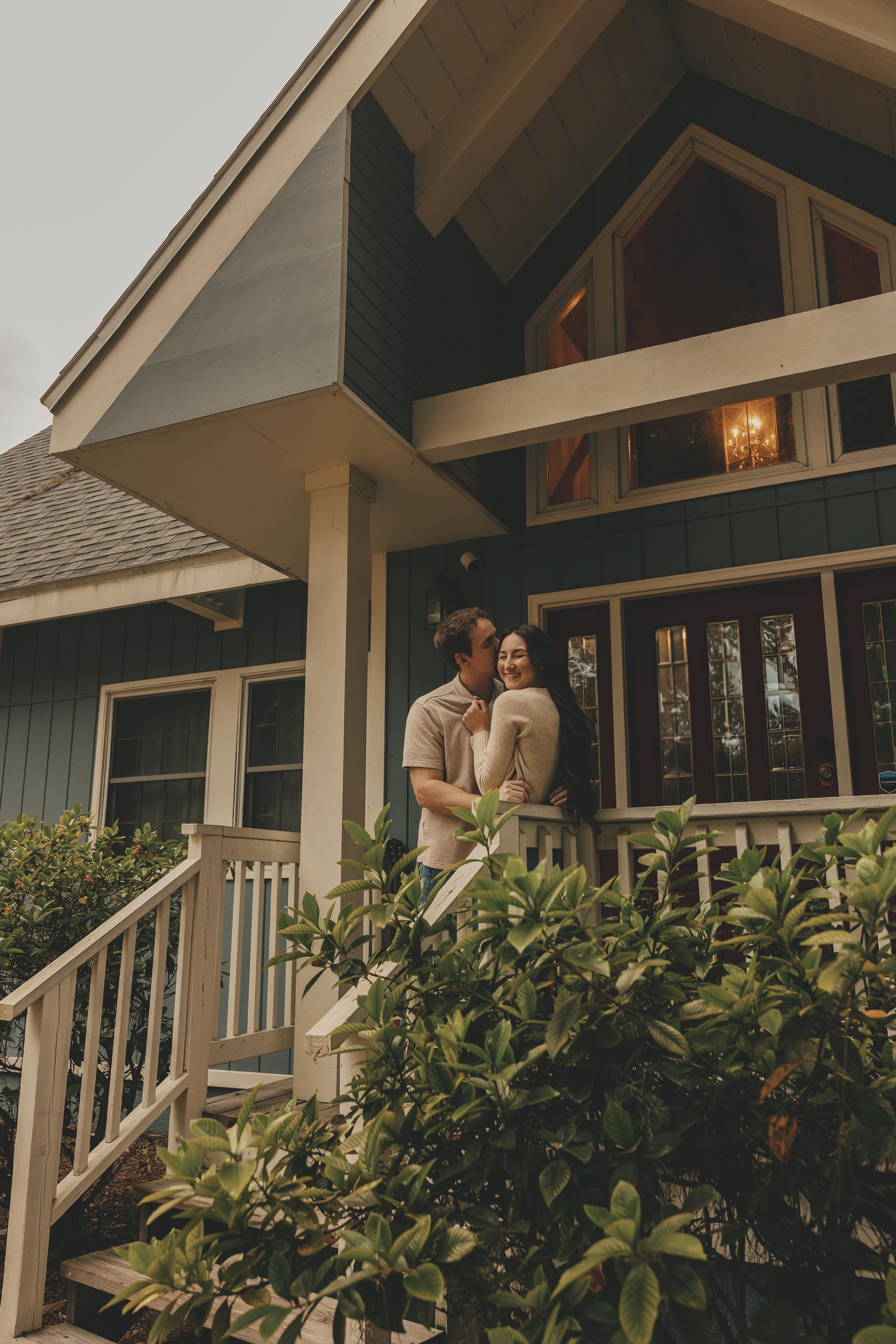 A couple embraces on the porch of a modern craftsman style house with white trim and lush greenery.
