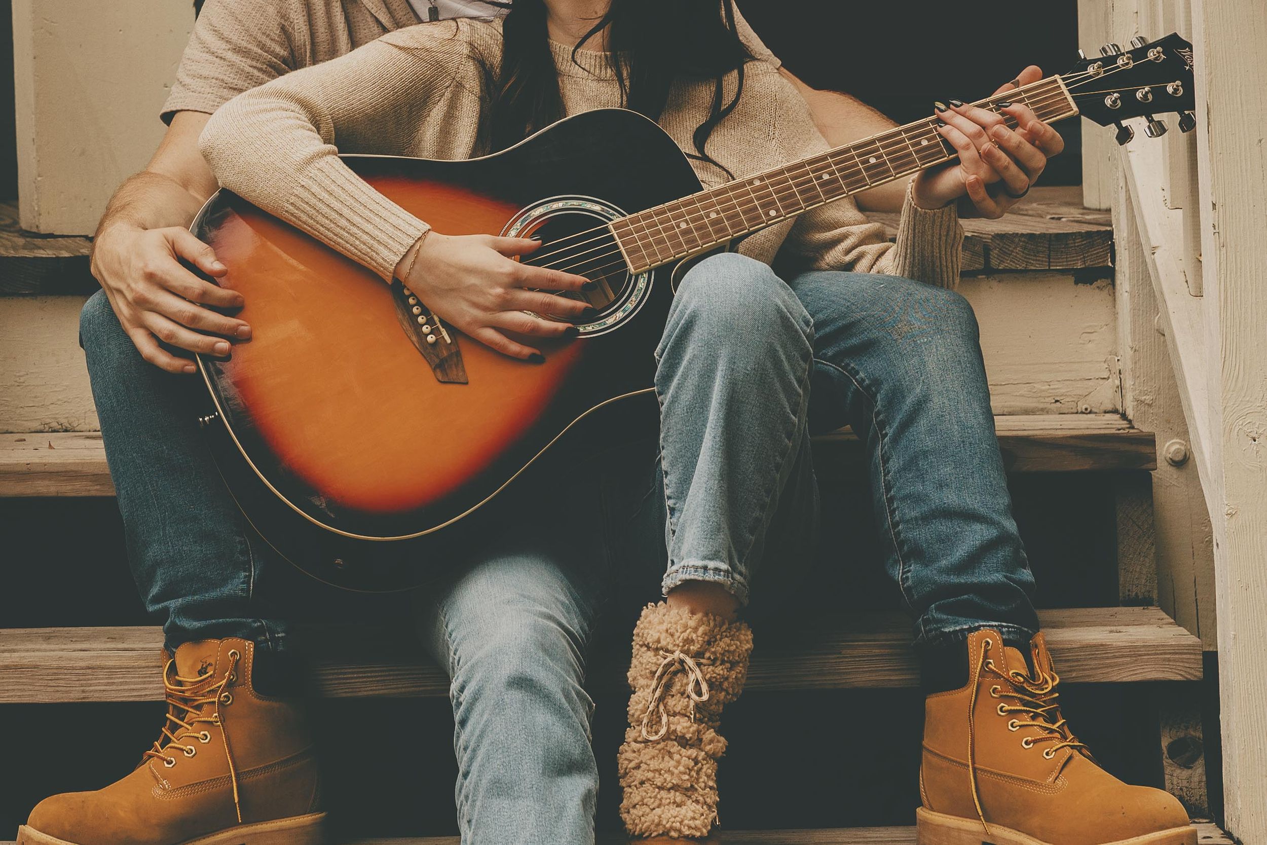 Close-up of hands playing acoustic guitar while sitting on wooden steps wearing brown boots.