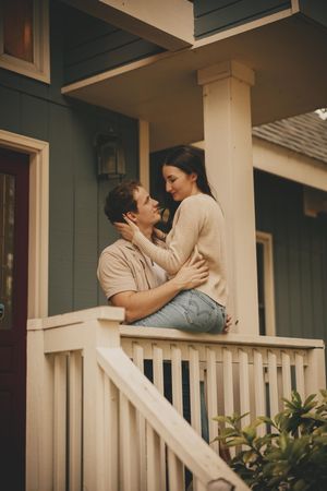 Romantic couple sharing intimate moment on front porch of house with white railing.