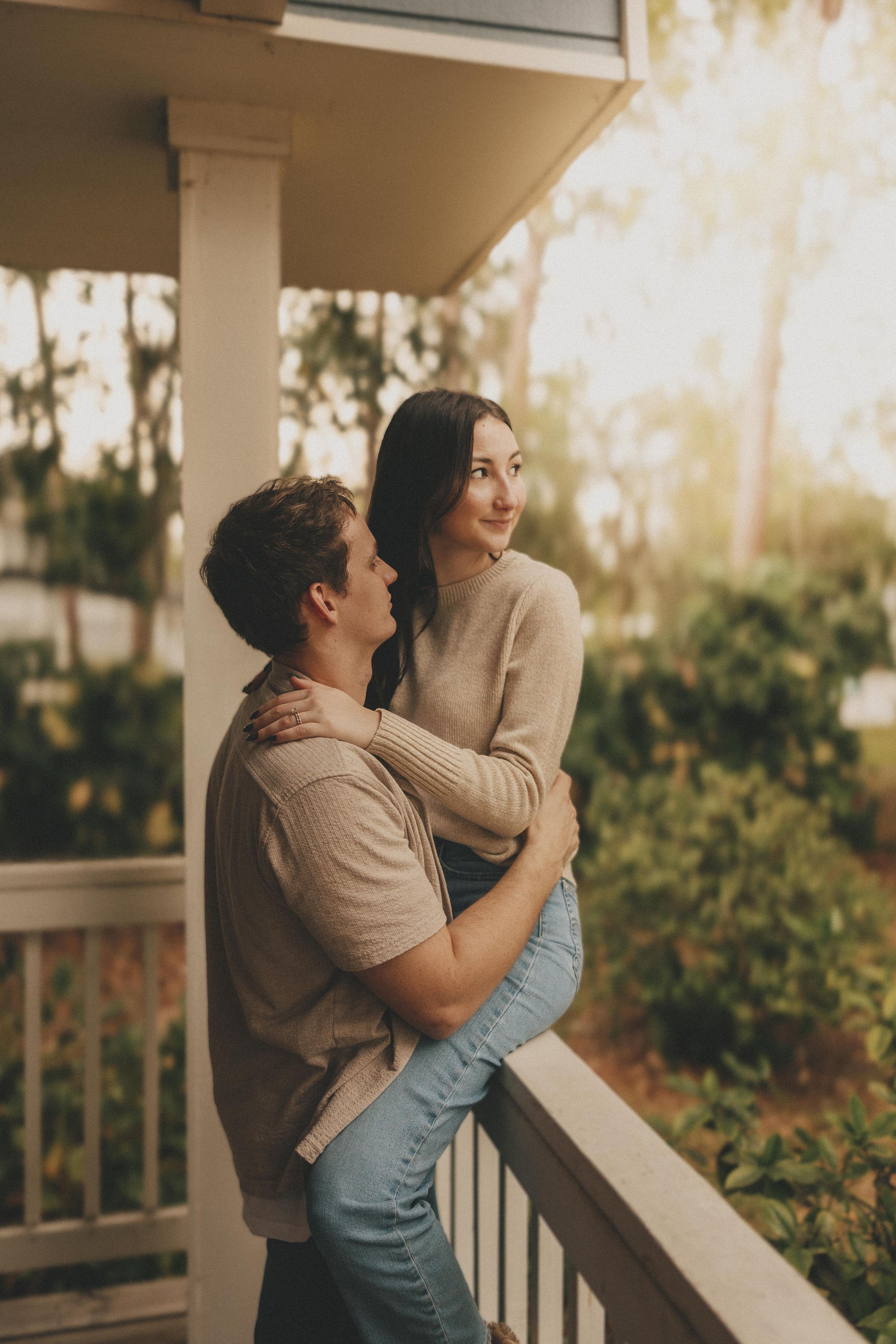 A romantic couple embraces on a porch at sunset with warm lighting and greenery in the background.