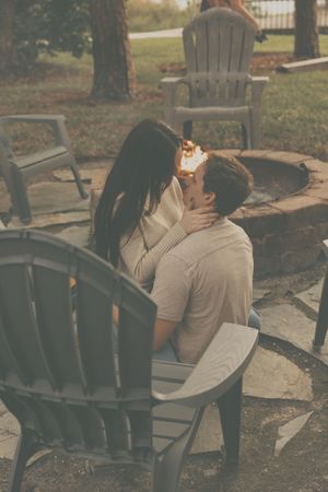A couple shares an intimate moment on outdoor chairs near a stone fire pit at dusk.